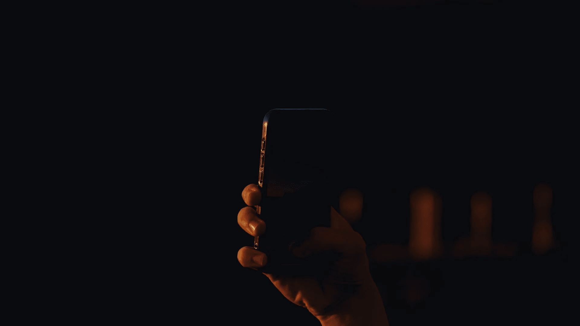 Hand holding a dark smartphone at night, with warm lights in the background.