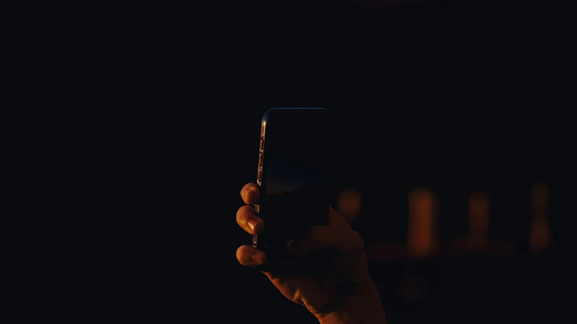 Hand holding a dark smartphone at night, with warm lights in the background.